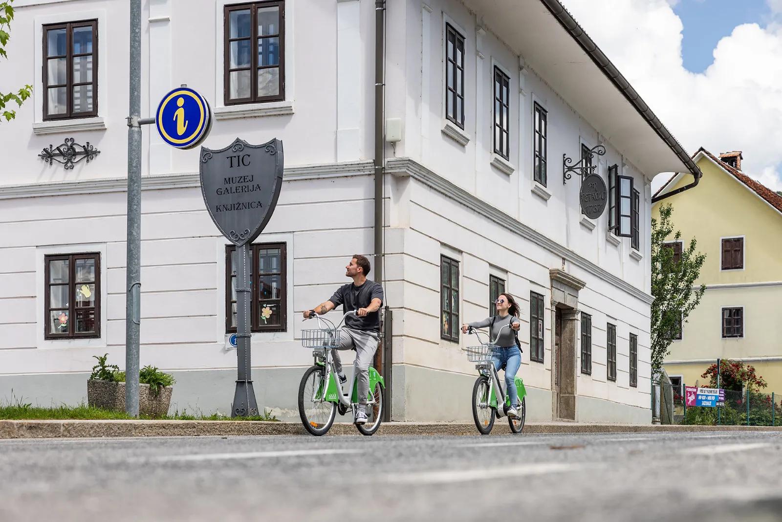 Kolesarja na zelenih kolesih s košarama se peljeta mimo bele hiše z oznako TIC, muzej, galerija, knjižnica.